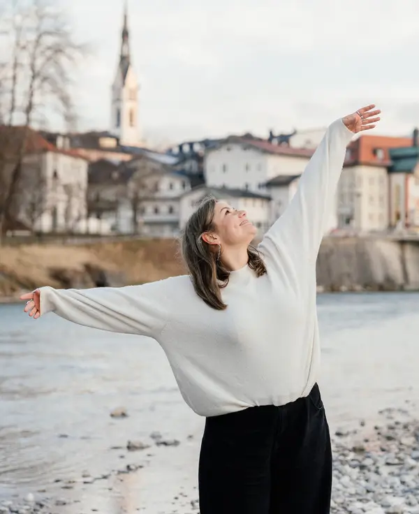 Jennifer Heng steht an einem Bachufer und hebt die Hände zum Himmel, schaut nach oben