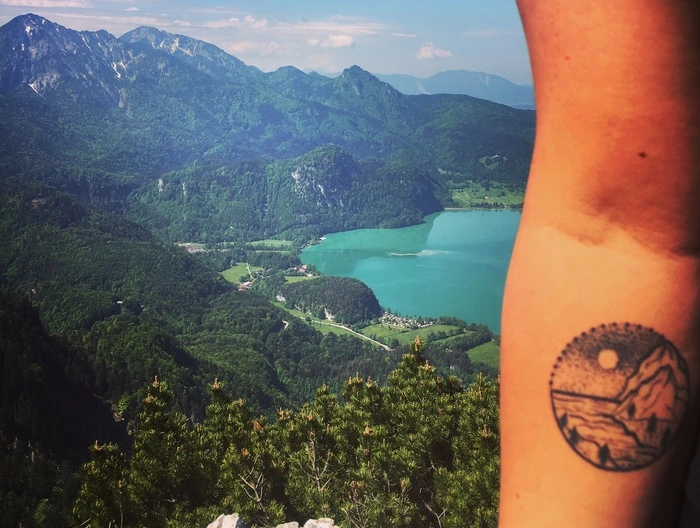 Der Arm mit dem "Berge und Meer"- Tattoo von Jennifer Heng vor einem Hintergrund mit Bergen und einem großen See